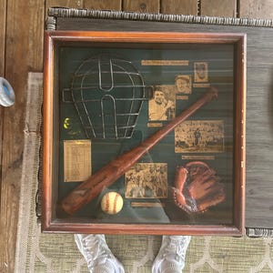 Pode incluir: Uma vitrine retangular de madeira contendo uma máscara de receptor de metal em estilo vintage, um taco de beisebol de madeira, uma bola de beisebol e uma luva de couro. A exibição apresenta fotos históricas e o texto The History of Baseball.