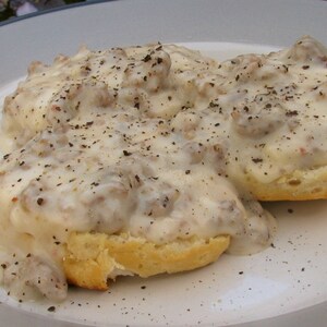 May include: A plate of biscuits and gravy. Two golden-brown biscuits are covered in a creamy white gravy with bits of sausage and black pepper. The dish is served on a white plate with a blue rim.