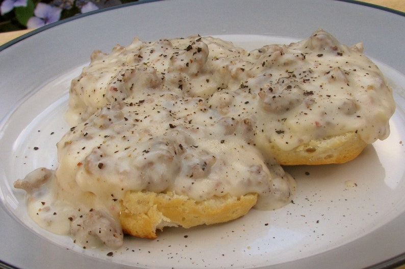 May include: A plate of biscuits and gravy. Two golden-brown biscuits are covered in a creamy white gravy with bits of sausage and black pepper. The dish is served on a white plate with a blue rim.