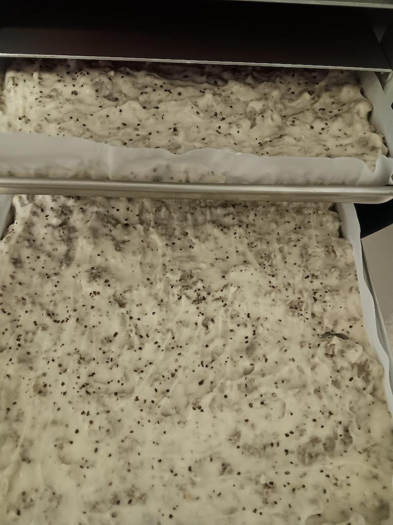 May include: Two rectangular baking pans filled with a light-colored batter speckled with dark seeds. The pans are in a dark oven, with one pan positioned above the other. The batter appears to be ready for baking.