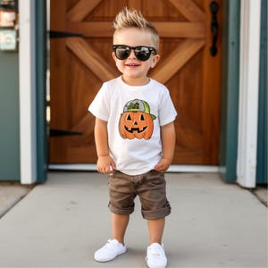 May include: A young child wearing a white t-shirt with a cartoon pumpkin graphic, brown shorts, white sneakers, and black sunglasses. The child is standing in front of a wooden door.
