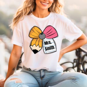 May include: White t-shirt with a yellow and pink bow made of a pencil and a bow. The bow has a black and white checkered pattern in the center. The text "Mrs. Smith" is written on a piece of paper with lines.