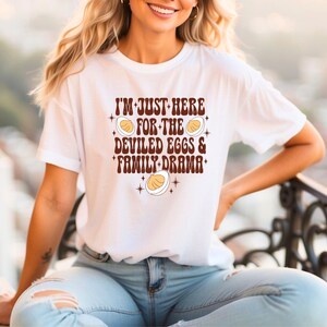 May include: White t-shirt with the text "I'M JUST HERE FOR THE DEVILED EGGS & FAMILY DRAMA" in brown lettering. The design includes illustrations of deviled eggs and stars. The shirt is a casual, everyday wear item.