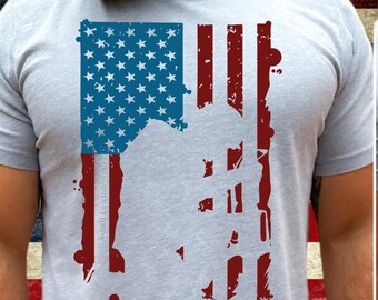 May include: A gray t-shirt with a distressed American flag design. The flag is red, white, and blue and has a silhouette of a person standing in front of it.