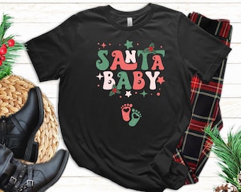 May include: Black t-shirt with a green and red "Santa Baby" graphic and a pair of green and red footprints. The shirt is on a white wooden surface with a plaid fabric and a green plant in the background.