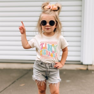 May include: A young girl wearing a white t-shirt with the text "Tiny Teenager" in rainbow colors and a checkered pattern. She is wearing denim shorts and round sunglasses.