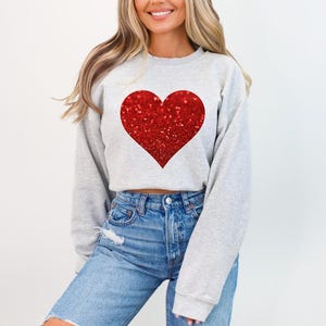May include: A gray sweatshirt with a large red glitter heart on the front. The sweatshirt is cropped and has long sleeves.