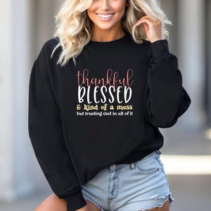 May include: Black sweatshirt with the words "thankful BLESSED & kind of a mess but trusting God in all of it" in pink and white script. The sweatshirt is paired with denim shorts.