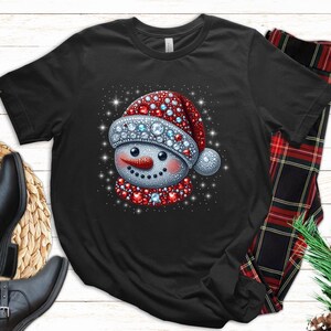 May include: Black t-shirt with a rhinestone snowman wearing a red and white Santa hat. The snowman has a red and white scarf and a white face with a red nose and black eyes.