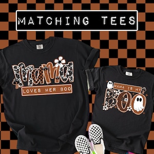 May include: Two black t-shirts with Halloween-themed designs. One shirt says "Mama Loves Her Boo" and the other says "Mama Is My Boo". The shirts have floral and mushroom accents, and a ghost graphic. The text "Matching Tees" is at the top.