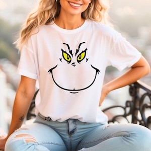 May include: White t-shirt featuring a graphic of a green-eyed, smiling face with black outlines. The design is centered on the front of the shirt. The shirt is a casual, short-sleeved style.