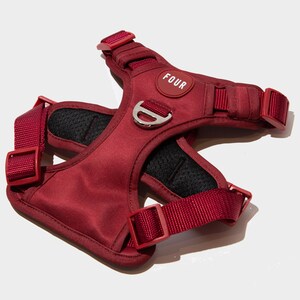 May include: A red dog harness with a black mesh lining and a silver buckle. The harness has a white label with the word "FOUR" printed on it.