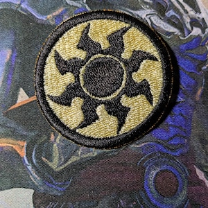 May include: A black and tan embroidered patch with a black sunburst design. The patch is circular and has a black border.
