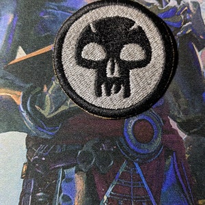 May include: A white and black embroidered skull patch with a black outline. The patch is circular and has a gray background.