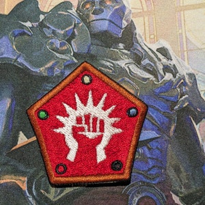 May include: A red, pentagon-shaped patch with a white symbol of a hand with a stylized sunburst behind it. The patch has a brown border and four colored circles around the symbol.