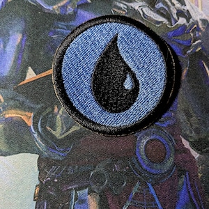 May include: A blue and black embroidered patch with a teardrop shape. The patch is circular and has a black border.