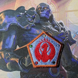 May include: A blue and red embroidered patch with a phoenix design. The patch is shaped like a pentagon and has a light blue background with a red phoenix in the center. The phoenix is surrounded by five small, colorful circles.