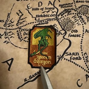 May include: A green dragon embroidered patch with the words "The Green Dragon" on a brown background. The patch is on a map of the Shire from the Lord of the Rings.