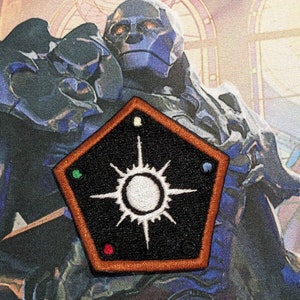May include: A black and orange pentagonal patch with a white starburst design and four colored dots around the starburst. The patch is embroidered with a white starburst and four colored dots.