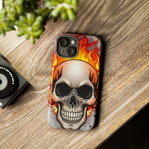 May include: A phone case with a skull design. The skull is surrounded by flames in shades of orange, yellow, and red. The phone case is made of a hard plastic material.