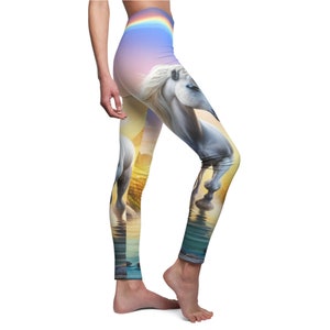 Puede incluir: Un par de leggings con un diseño de arcoíris colorido en la cintura y un unicornio blanco con una crin y cola de arcoíris de pie en un cuerpo de agua. El unicornio se refleja en el agua.