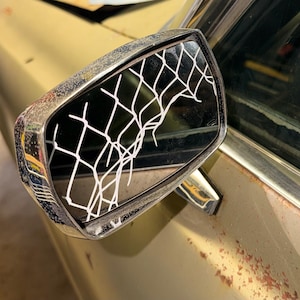 Könnte beinhalten: Nahaufnahme eines Autoseitenspiegels mit einem weißen Netzdesign auf dem schwarzen Glas. Der Spiegel hat einen Chromrahmen und ist an einer goldfarbenen Karosserie befestigt. Das Auto weist Gebrauchsspuren auf.
