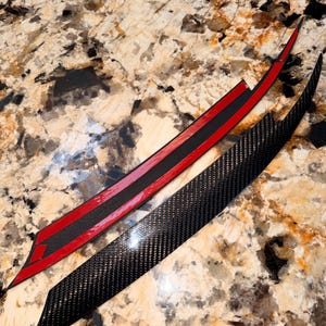 May include: Two long, slender, curved automotive trim pieces featuring a black carbon fibre weave pattern. One piece displays its smooth, textured carbon fibre surface, while the other shows its underside with a strip of red adhesive backing tape.