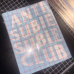 以下が含まれることがあります： 白いブロック体の「ANTI SUBTLE CLUB」という文字が入った水色のステッカー。ステッカーは、赤い格子模様のカッティングマットとペーパーカッターの上にあります。