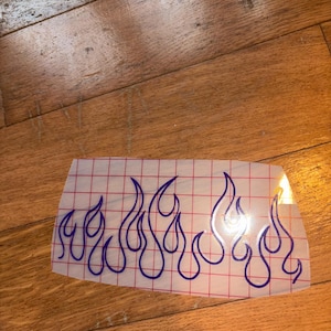 May include: A clear vinyl decal featuring a blue flame design. The flames are outlined in blue and arranged in a horizontal pattern. The decal is on a grid-patterned backing, suitable for application on various surfaces.