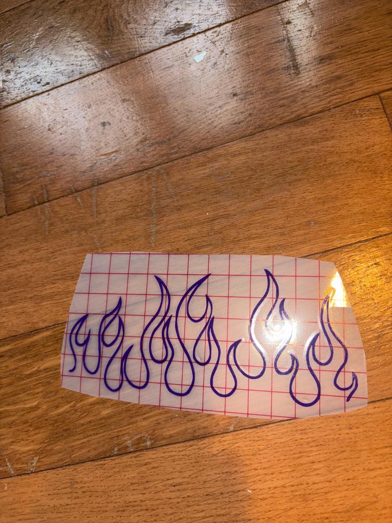 May include: A clear vinyl decal featuring a blue flame design. The flames are outlined in blue and arranged in a horizontal pattern. The decal is on a grid-patterned backing, suitable for application on various surfaces.