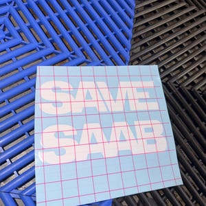 May include: A square light blue sticker featuring a pink grid pattern and the white text SAVE SAAB in a bold, blocky font. The sticker is positioned against a background of interlocking blue and black plastic floor tiles with a geometric design.