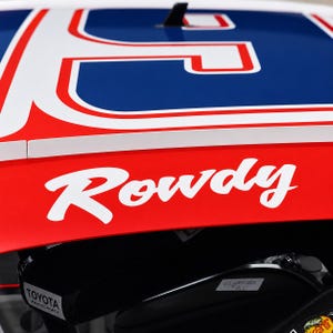 Pode incluir: Um painel de teto de carro de corrida vermelho, branco e azul com a palavra Rowdy em letra cursiva branca. O painel exibe um grande número azul com contorno branco e vermelho, uma antena preta e um logotipo da Toyota Racing em uma peça preta.