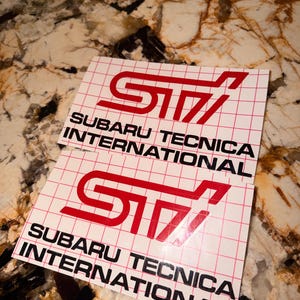 May include: Two white stickers featuring the red STI logo and the text "SUBARU TECNICA INTERNATIONAL" in black. The stickers have a red grid pattern.