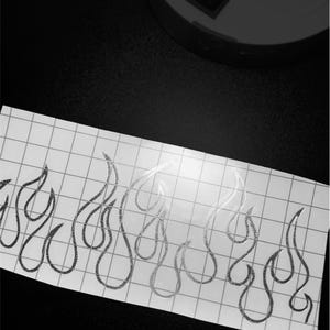 May include: A sheet of white grid paper with a silver glitter flame design. The flames are stylized and outlined. The background is black, with a white circular object and a switch visible.
