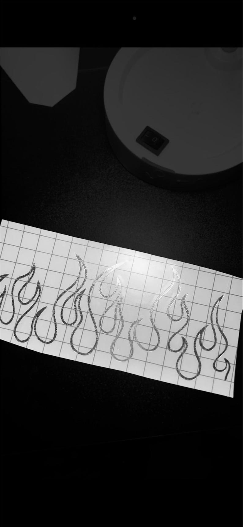 May include: A sheet of white grid paper with a silver glitter flame design. The flames are stylized and outlined. The background is black, with a white circular object and a switch visible.
