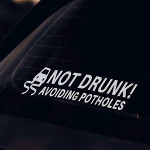 Puede incluir: Adhesivo de vinilo blanco para coche con el texto "NOT DRUNK! AVOIDING POTHOLES". Incluye un dibujo de un coche y una línea sinuosa. El texto es en negrita y sin serifa.
