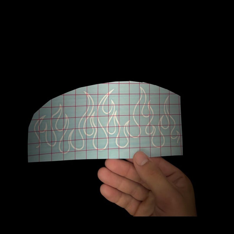 May include: White flame decal on a light blue grid background. The decal features a series of stylized flames. The decal is held by a hand against a black background. The flames are a popular design element for vehicles and other items.