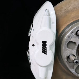May include: A white brake caliper with the black "M" logo, mounted next to a rotor. The caliper is a bright white color, with a smooth finish. The rotor is a metallic gold color.