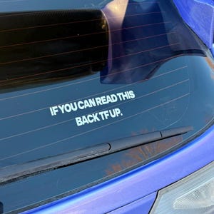 Può includere: Un adesivo bianco sul lunotto posteriore di un'auto con la scritta "IF YOU CAN READ THIS BACK TF UP." L'adesivo è centrato sotto il tergicristallo posteriore. L'auto è blu.