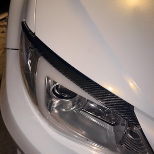 Könnte beinhalten: Nahaufnahme eines weißen Autoscheinwerfers mit einem schwarzen Kohlefaserakzent. Der Scheinwerfer ist klar und hat mehrere interne Reflektoren. Die Karosserie des Autos ist weiß und hat eine glänzende Oberfläche.