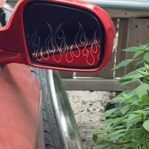 May include: Red side mirror with a flame design. The flames are outlined in white against a black background. The mirror is attached to a red vehicle, with a tire visible in the foreground.