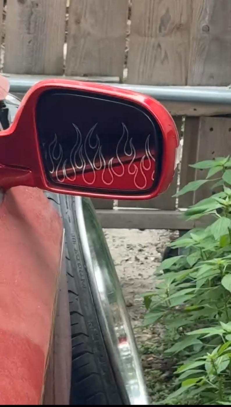 May include: Red side mirror with a flame design. The flames are outlined in white against a black background. The mirror is attached to a red vehicle, with a tire visible in the foreground.