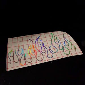 May include: A sheet of flame-shaped stickers with a holographic, rainbow-colored effect. The flames are outlined in a dark color and arranged in a row. The stickers are on a white sheet with a red grid pattern.