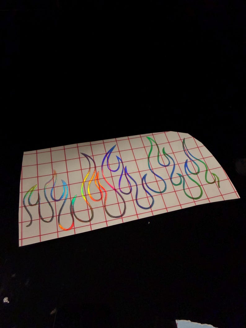 May include: A sheet of flame-shaped stickers with a holographic, rainbow-colored effect. The flames are outlined in a dark color and arranged in a row. The stickers are on a white sheet with a red grid pattern.