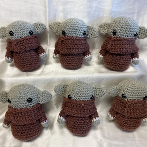 May include: Six crocheted plush toys in the shape of a fictional character with large ears, brown eyes, and a brown sweater. The toys are arranged in two rows of three.