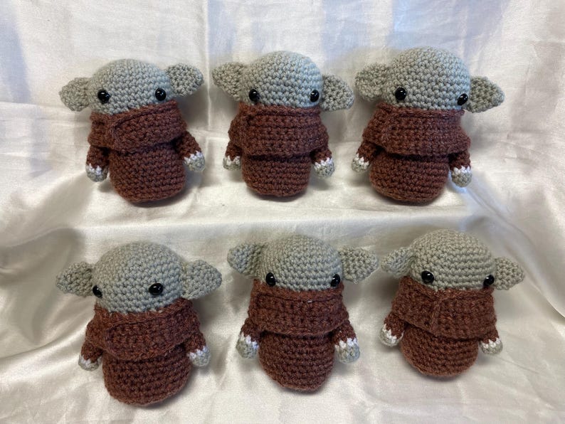 May include: Six crocheted plush toys in the shape of a fictional character with large ears, brown eyes, and a brown sweater. The toys are arranged in two rows of three.