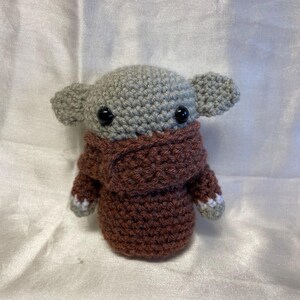 May include: A crocheted plush toy in the shape of a small, gray alien with large eyes. The alien is wearing a brown crocheted sweater with white accents.
