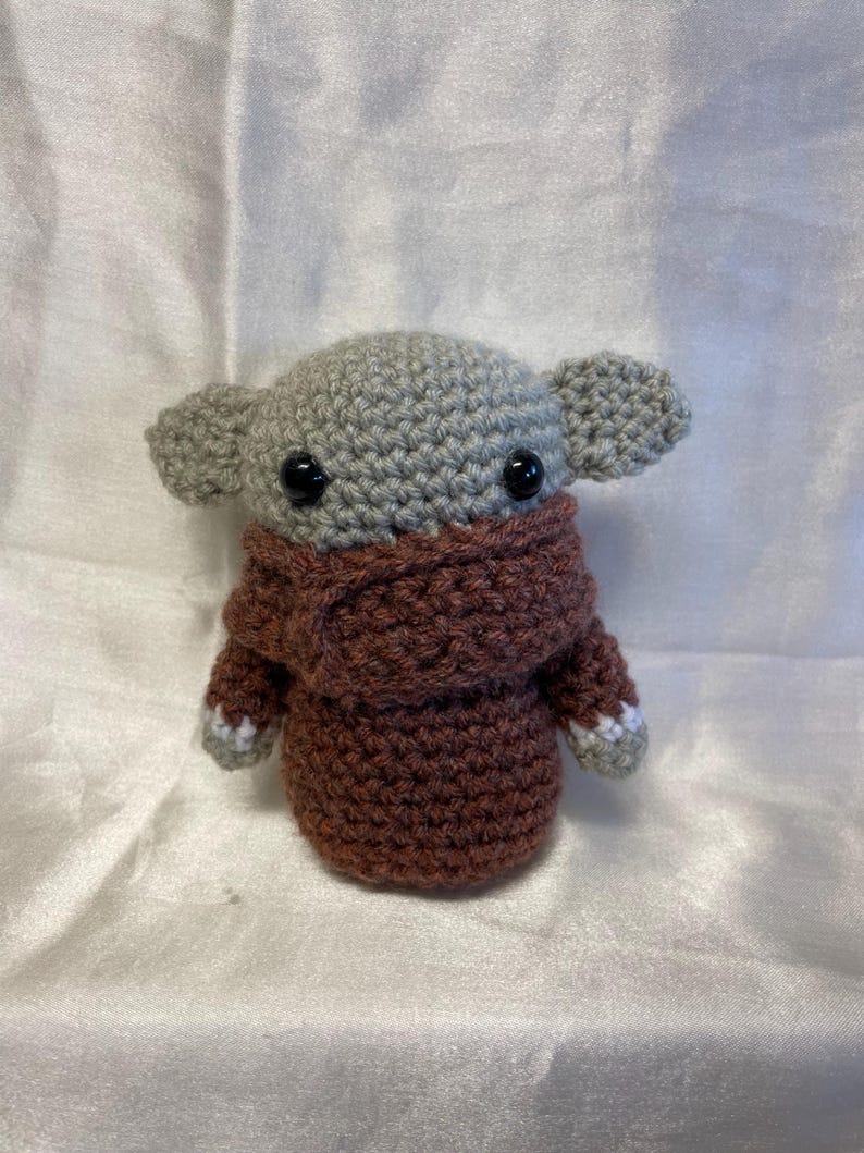 May include: A crocheted plush toy in the shape of a small, gray alien with large eyes. The alien is wearing a brown crocheted sweater with white accents.