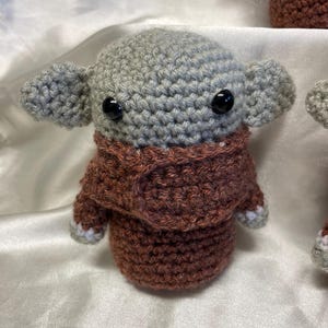 May include: A crocheted plush toy with a gray head and brown body. The toy has large black eyes and a brown crocheted scarf.