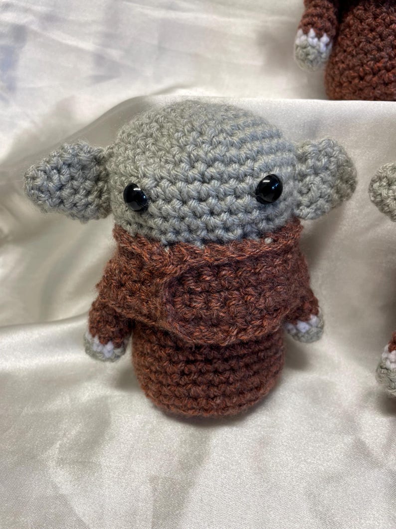 May include: A crocheted plush toy with a gray head and brown body. The toy has large black eyes and a brown crocheted scarf.